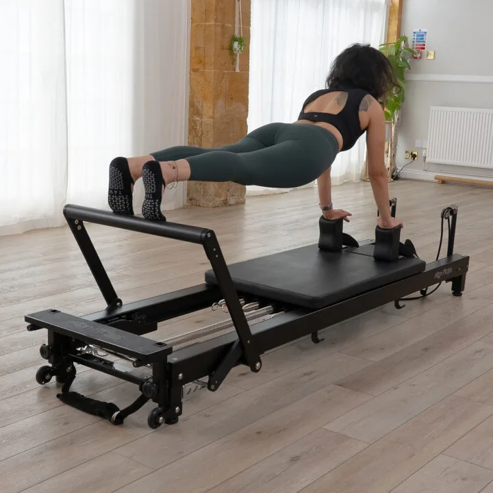 Woman performing plank exercise on black Pilates reformer in bright studio space.